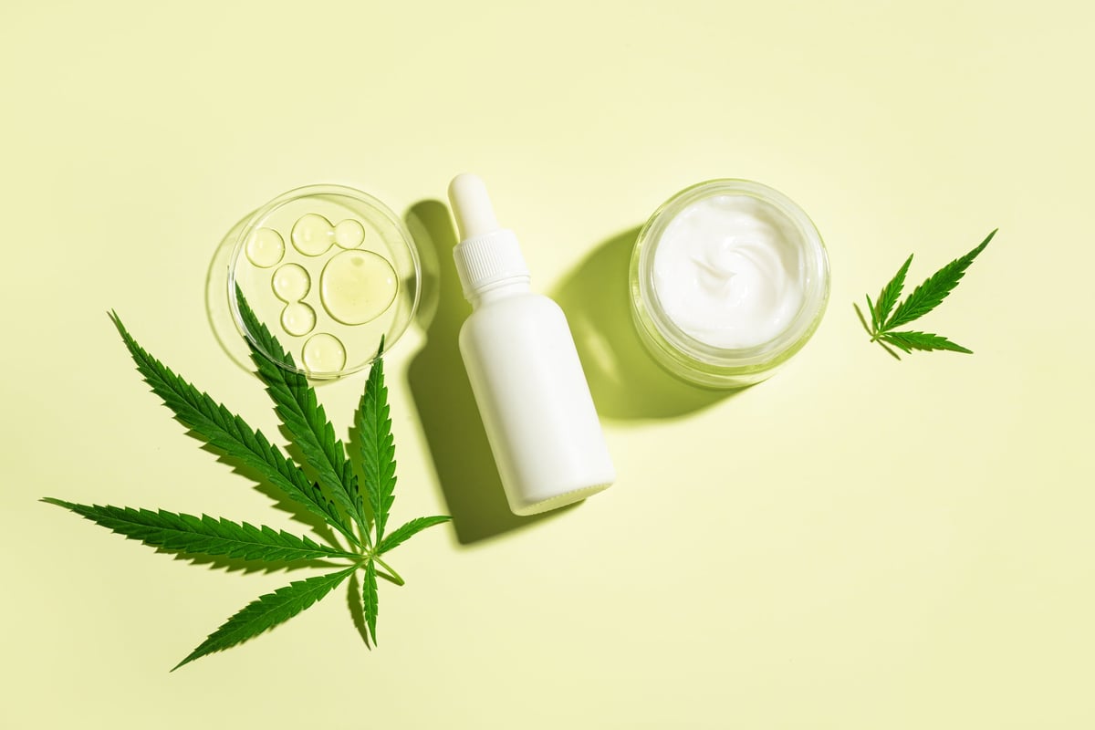 CBD oil, hemp leaves, and cosmetic products on light yellow background